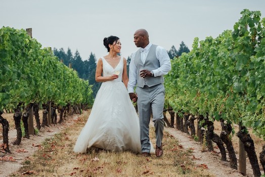 Bride and groom walk hand in hand down a vineyard row, smiling at each other with green vines on either side.