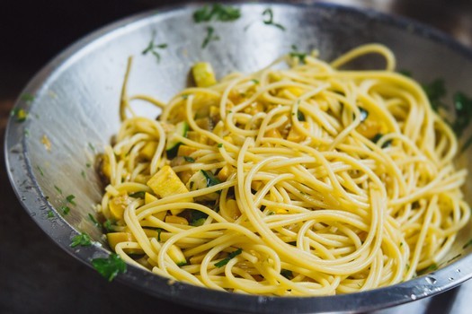 Close-up of freshly cooked spaghetti tossed with herbs and vegetables in a large stainless steel bowl.