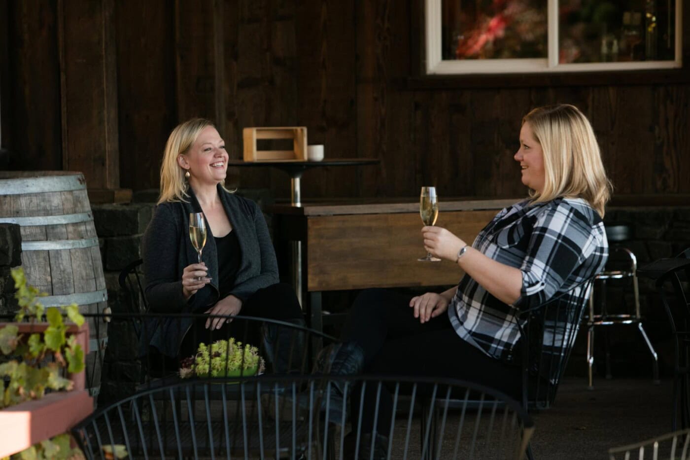Two women seated outside near wine barrels, smiling and toasting with glasses of sparkling wine.