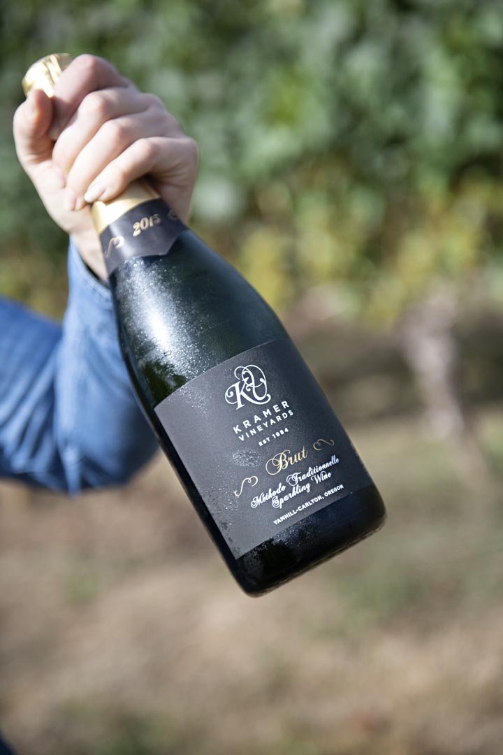 Hand holding a chilled bottle of Kramer Vineyards Brut sparkling wine outdoors, with vineyard greenery in the background.